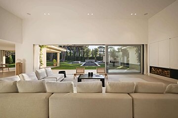 A bright, modern living room with cream sofas and expansive glass doors opening to a manicured lawn, swimming pool, and patio area.