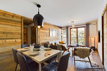A spacious, warm chalet interior with wood-paneled walls, a set dining table with fondue, and a cozy living area featuring fur-covered chairs by large windows.