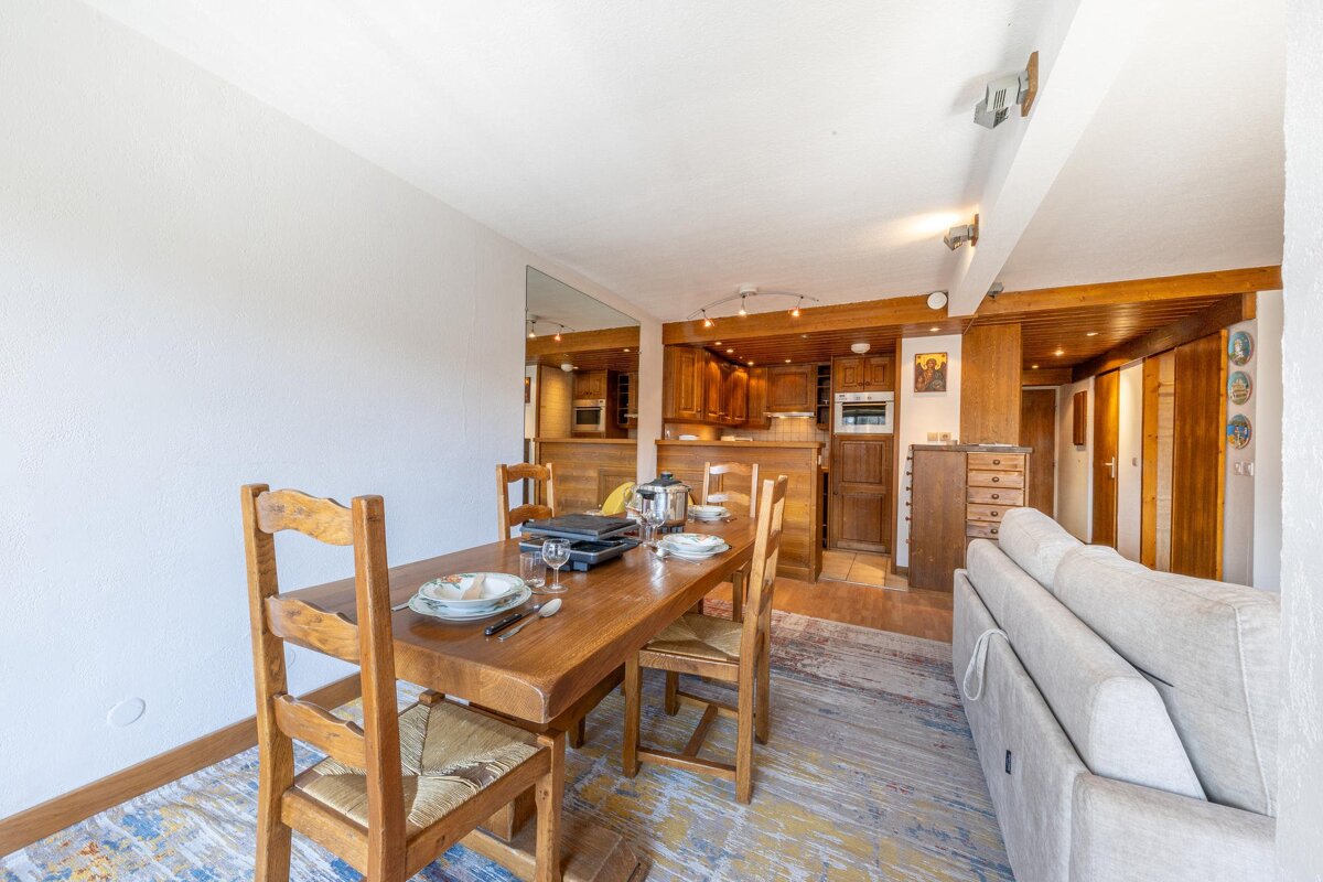 A cozy, open-concept room with a wooden dining table, kitchen, and sofa. Rustic wood accents and a raclette grill on the table complete the scene.