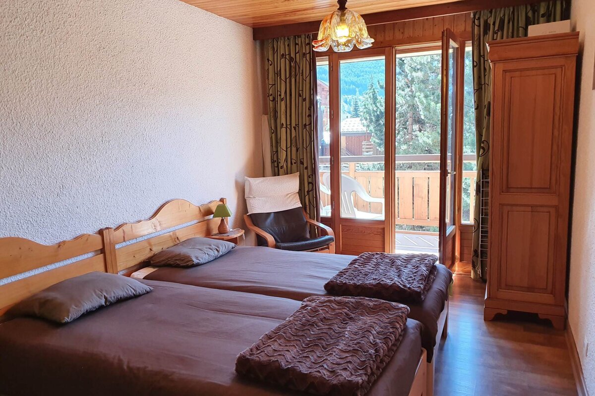 A cozy twin bedroom with wooden beds and furniture, featuring a balcony with mountain and forest views. Wood ceiling and textured walls.