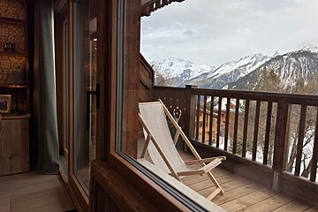 A wooden chalet balcony with a white deck chair offers a stunning view of snow-capped mountains under a cloudy sky. The cozy interior is visible through glass.