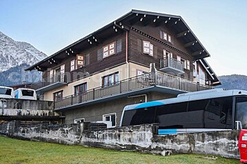 A multi-story alpine building with balconies, modern shuttle vehicles stored on concrete platforms, and snow-capped mountains in the background.