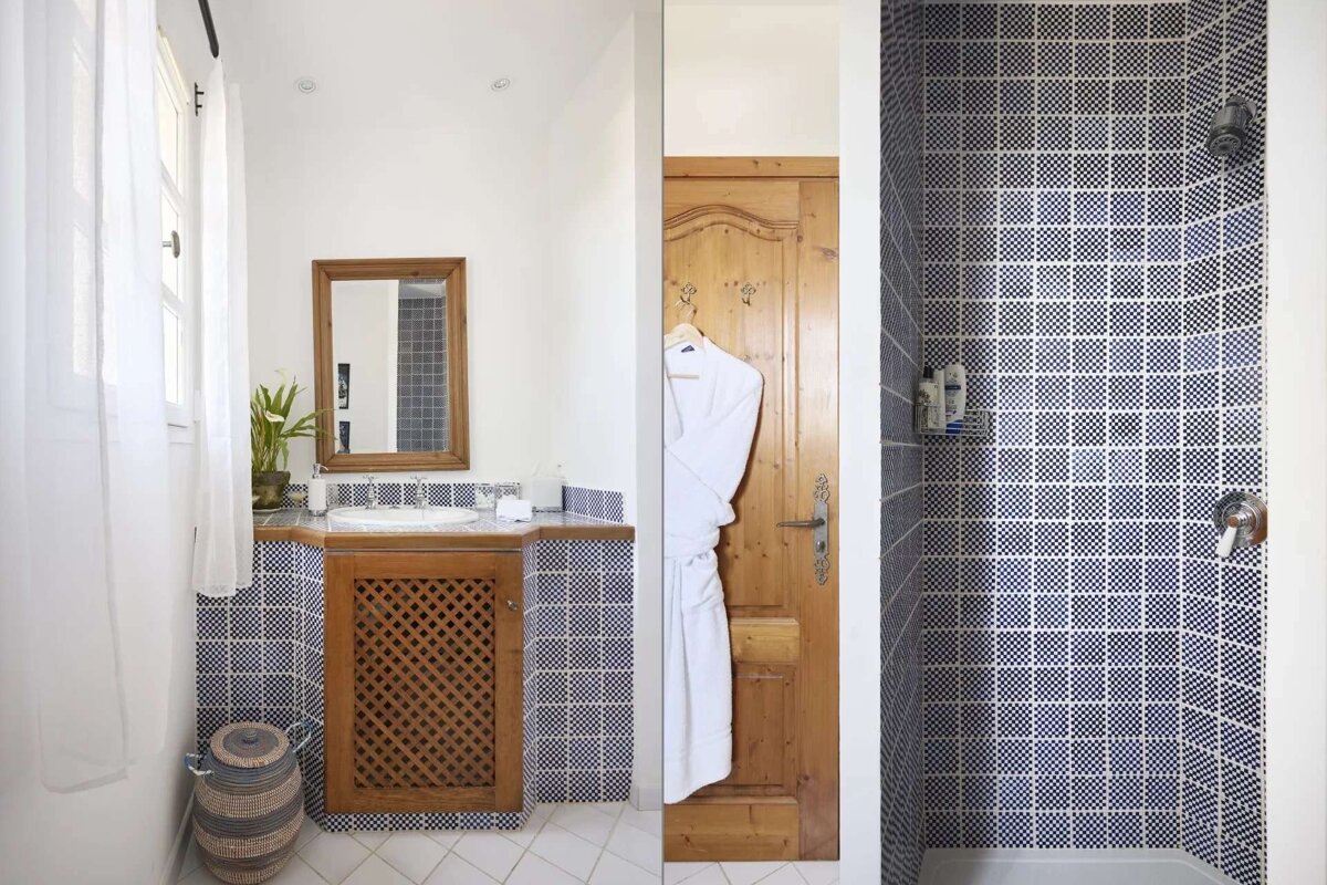 A bright bathroom features a wooden vanity with a mirror, blue and white tiled walls, and a shower area. A white bathrobe hangs on a wooden door.