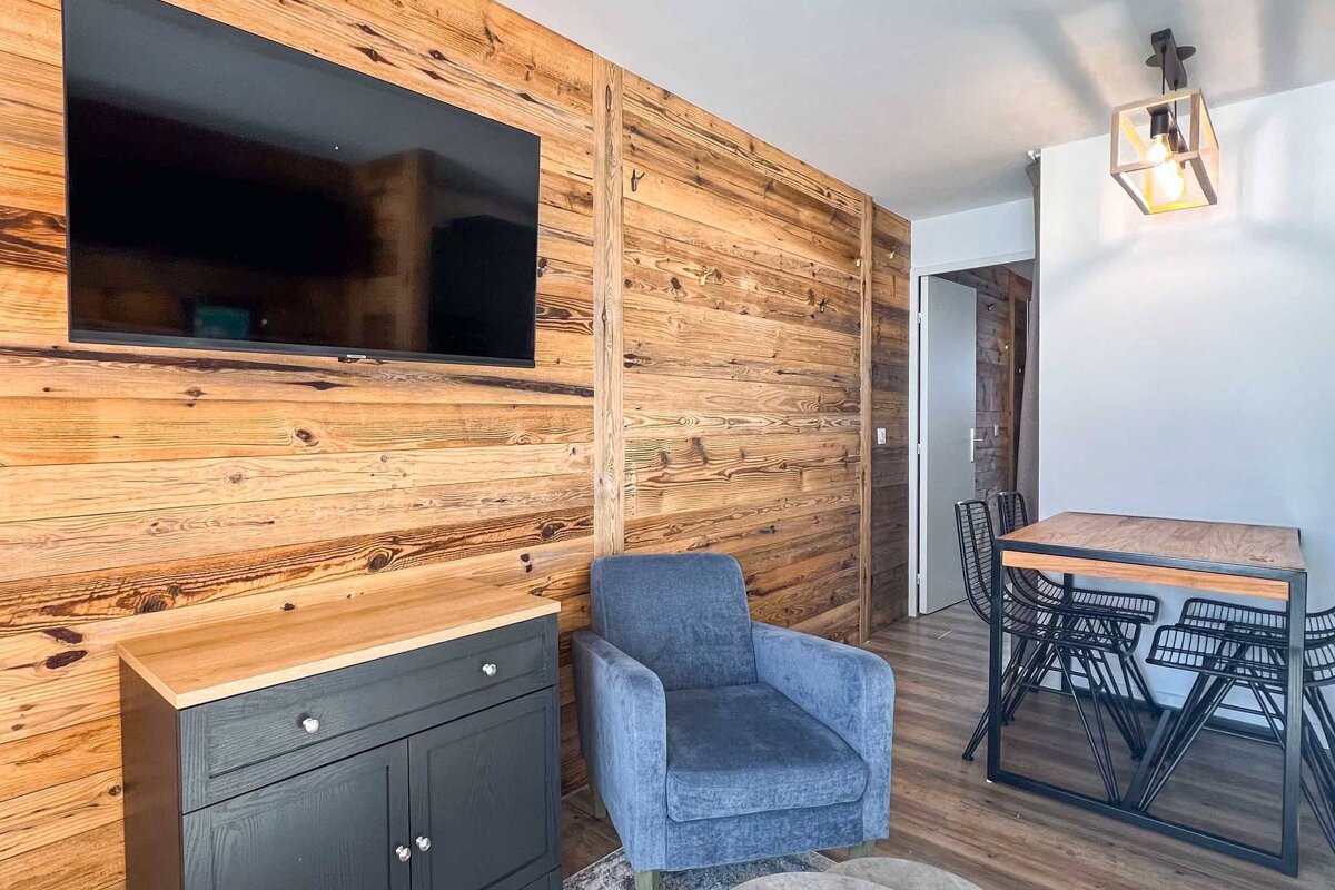 A modern rustic living area featuring a prominent wood-paneled wall with a large TV, a blue armchair, and a dining table with black chairs.