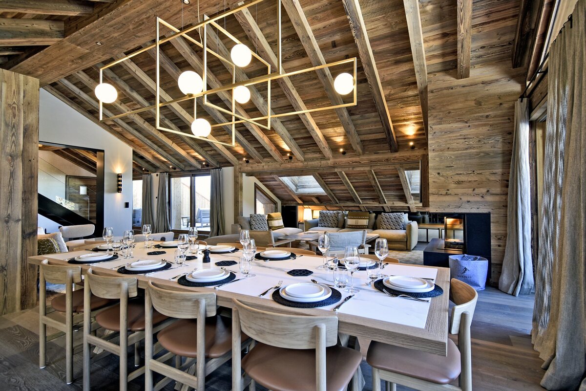 A luxurious open-plan chalet interior with a large dining table set, a cozy living area, exposed wooden beams, and a modern chandelier.