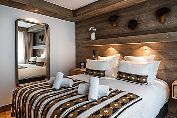 Rustic-chic bedroom featuring a wooden accent wall and ceiling, white bedding with a patterned throw, and a large mirror. Warm lights illuminate the bed.
