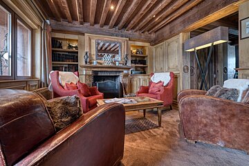 Cozy chalet-style living room with exposed wooden beams, a fireplace, leather and red chairs, bookshelves, and a warm ambiance.