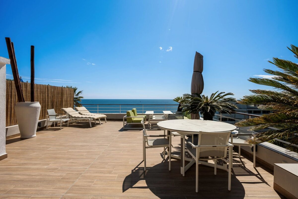 Sunny outdoor terrace with stunning ocean views, featuring lounge chairs, dining furniture, and palm trees under a clear blue sky.