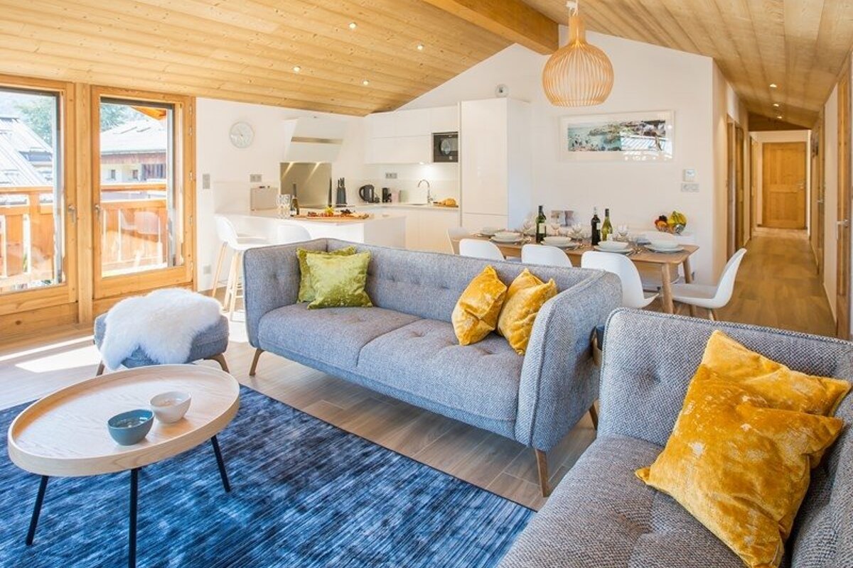 A cozy, modern open-plan living room with gray sofas, yellow pillows, a blue rug, wooden ceilings, and an adjoining kitchen.