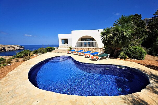 A white villa with a kidney-shaped swimming pool overlooks the blue ocean and coastline under a clear sky. Lounge chairs sit by the pool, surrounded by lush palms.