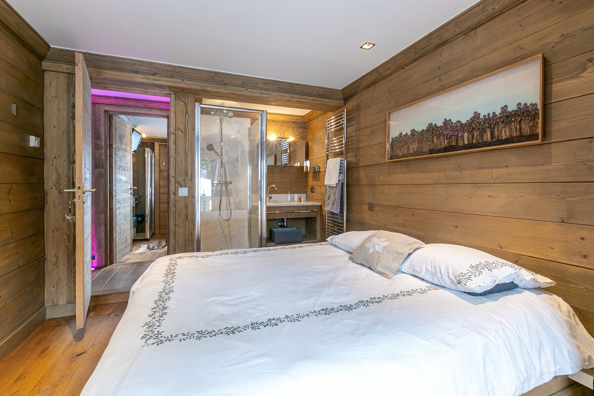 Cozy chalet bedroom with wood paneling, white bed, and an open-concept en-suite bathroom featuring a glass shower and sink.