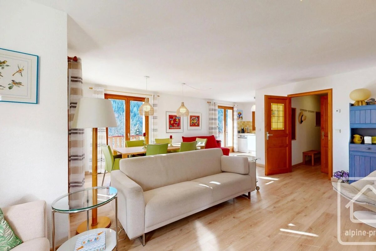 Sunlit, open-plan living and dining area with a cream sofa, wooden floors, and a dining table with green chairs.