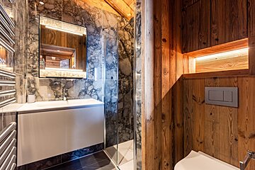 Modern rustic bathroom featuring marble walls, a glass shower, a white vanity with an illuminated mirror, and warm wooden paneling.