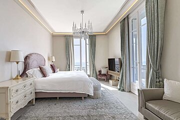 A luxurious, elegant bedroom features a large bed, chandelier, tall balcony windows with an ocean view, and soft pastel decor.