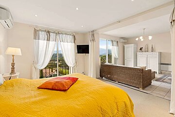 A bright, open-plan room featuring a yellow bed, a wicker sofa in a connected living area, and large windows with a scenic mountain view.