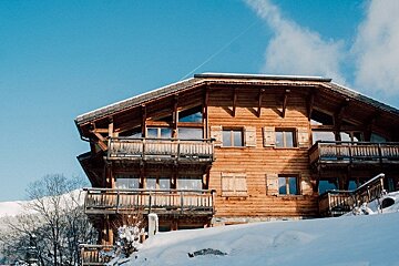 A large wooden alpine chalet on a snow-covered slope under a clear blue sky.