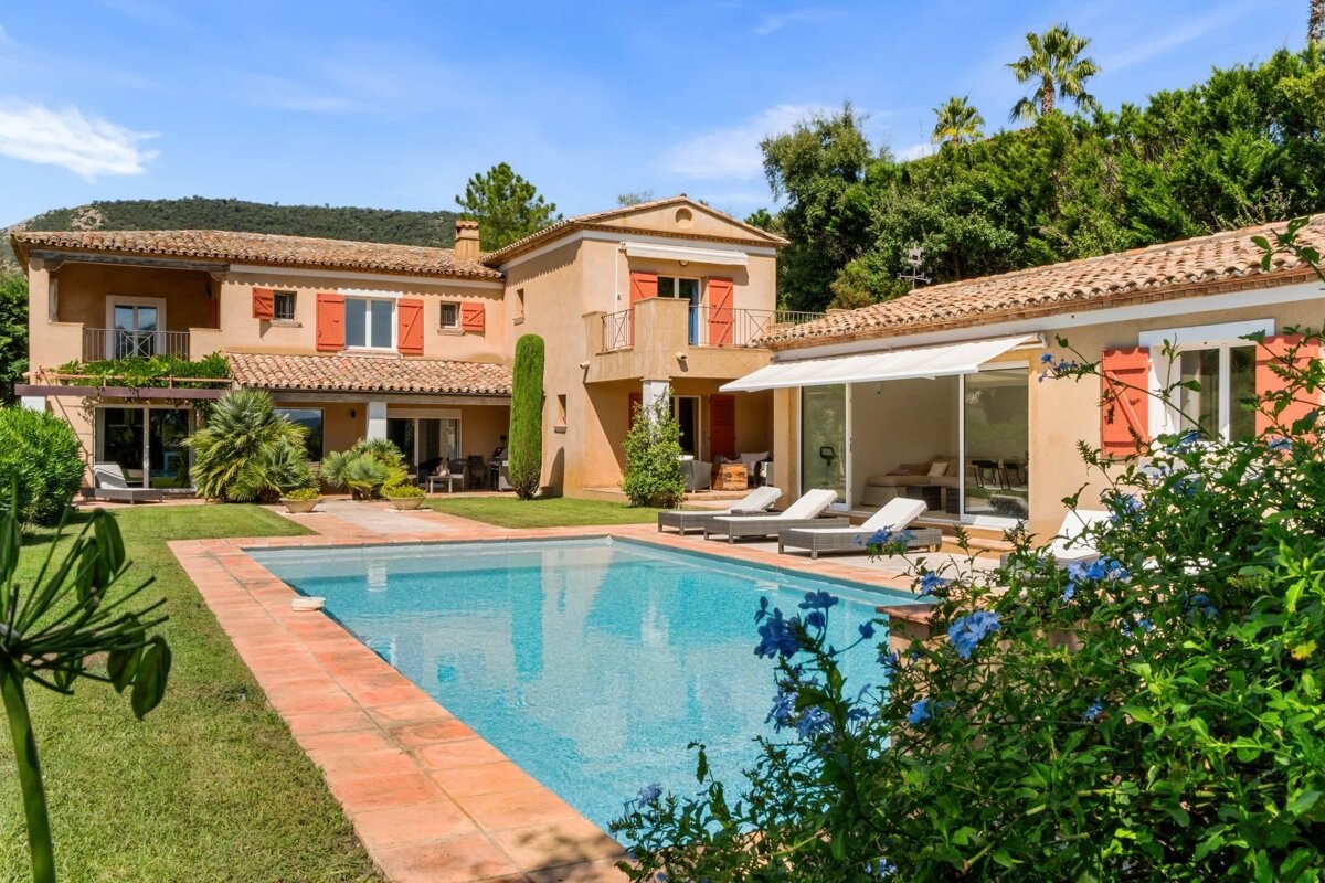 A beautiful Mediterranean villa with a sparkling swimming pool, green lawns, and sun loungers under a clear blue sky.