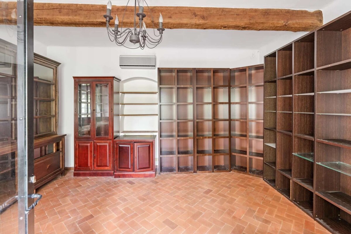 An empty room with terracotta tile floor, white walls, wooden beam ceiling, chandelier, and a mix of built-in and freestanding empty wooden bookshelves/cabinets.