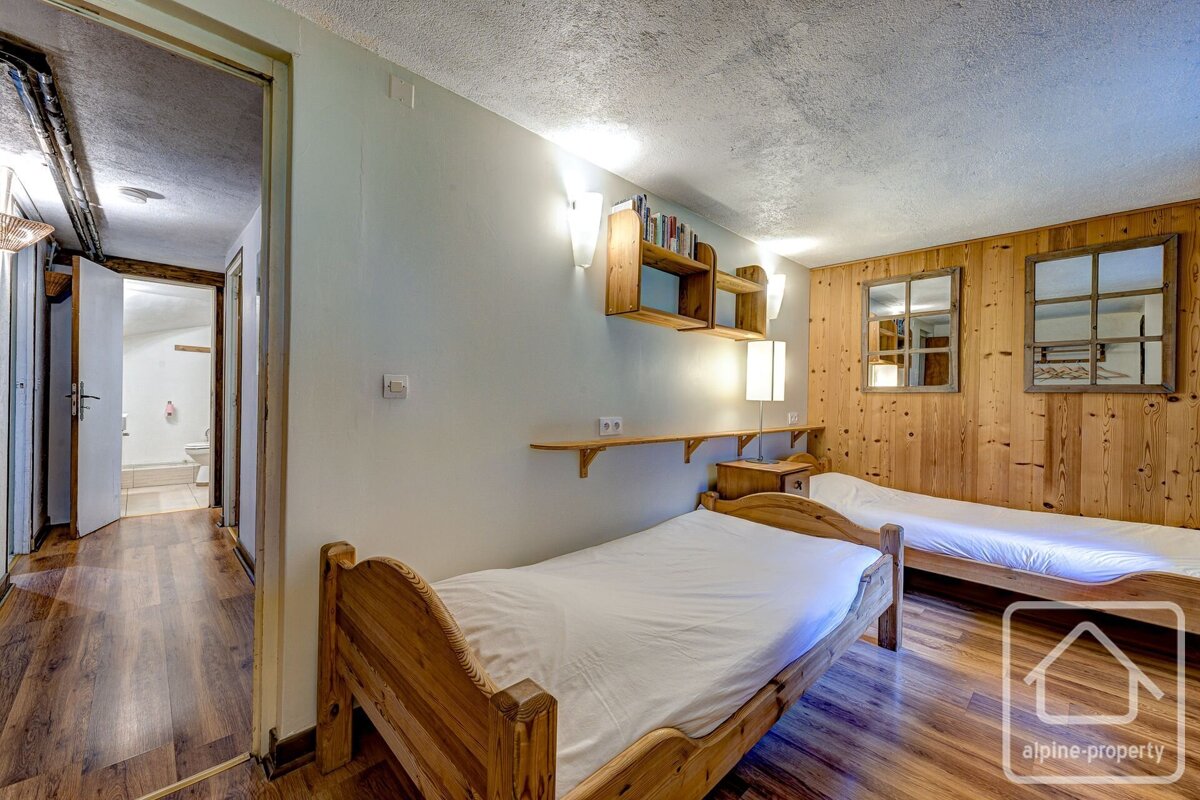 A cozy bedroom features two single beds with wooden frames, a wood-paneled wall, a light wall with shelves, wooden flooring, and a view into a bathroom.