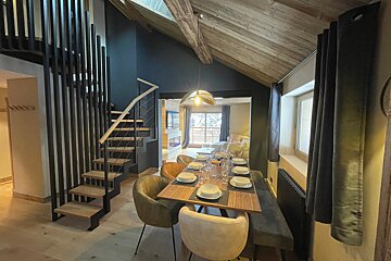 A modern chalet dining area features a set table, a stylish wooden staircase, and a vaulted ceiling with beams, opening to a cozy living space.