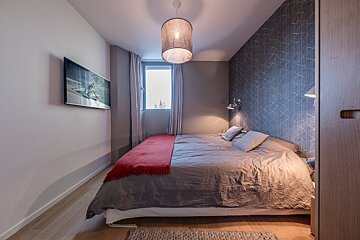 A modern bedroom with a grey bed, red throw, wall-mounted TV showing a skier, patterned wallpaper, and a window.