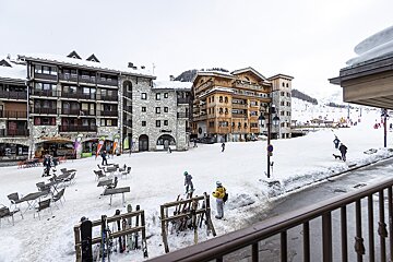 A snowy ski resort village square with stone and wooden buildings, outdoor seating, ski/snowboard racks, and people milling about.