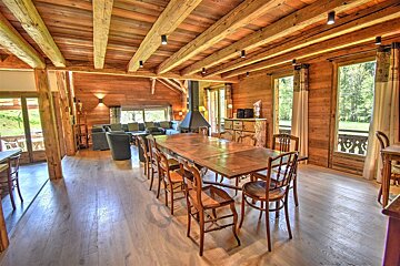 A rustic wooden chalet interior featuring a long dining table, exposed ceiling beams, and a cozy living area in the background.