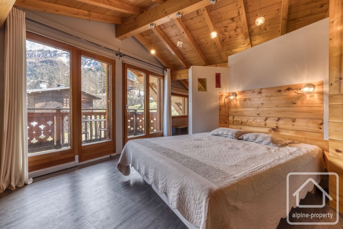 Cozy wood-paneled bedroom with large windows, a balcony, and mountain views, featuring a wooden beamed ceiling and headboard.