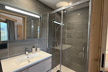 Modern bathroom with grey tiled walls, a glass shower stall, and a white sink vanity beneath an illuminated mirror.