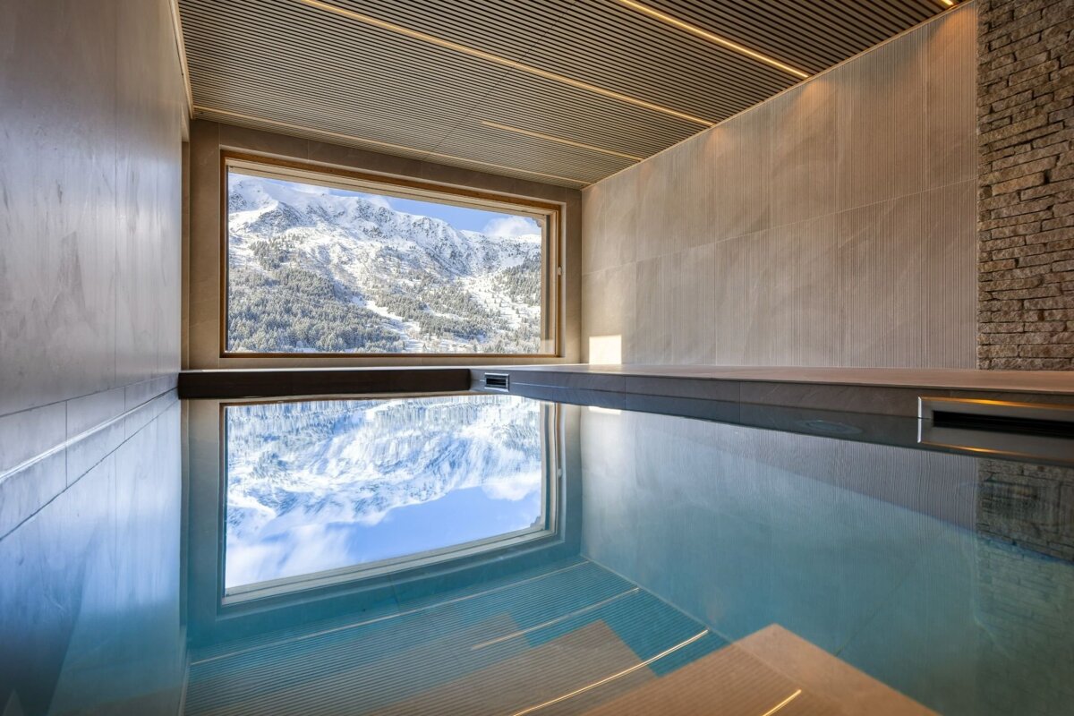 An indoor pool with a large window overlooking snow-covered mountains, beautifully reflected in the still water.