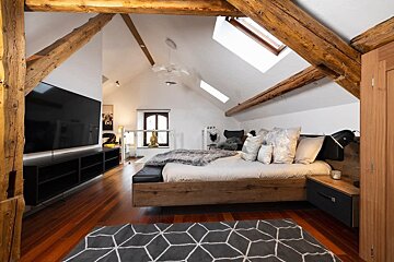 A modern attic bedroom features exposed wooden beams, vaulted ceilings with skylights, a large bed, TV, warm wooden floor, and a geometric rug.