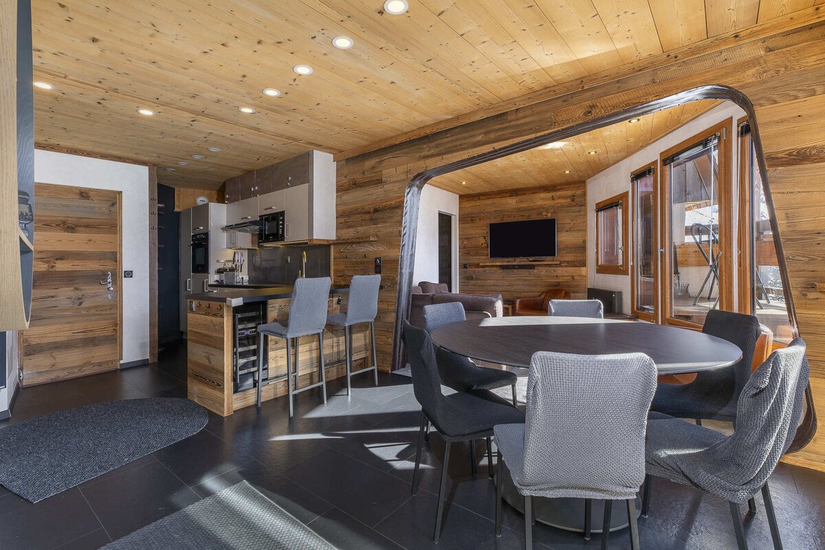 Modern chalet interior with warm wood paneled walls, a dining table with grey chairs, and an adjacent kitchen bar area.