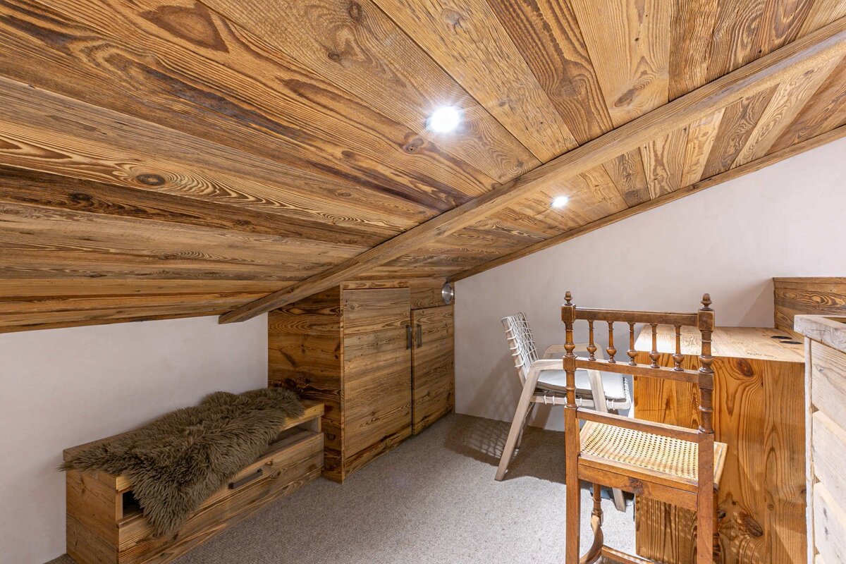 A cozy attic room with a slanted wooden plank ceiling, white walls, and light brown carpet. Wooden furniture, including a fur-covered bench, cabinets, and chairs, fills the space.