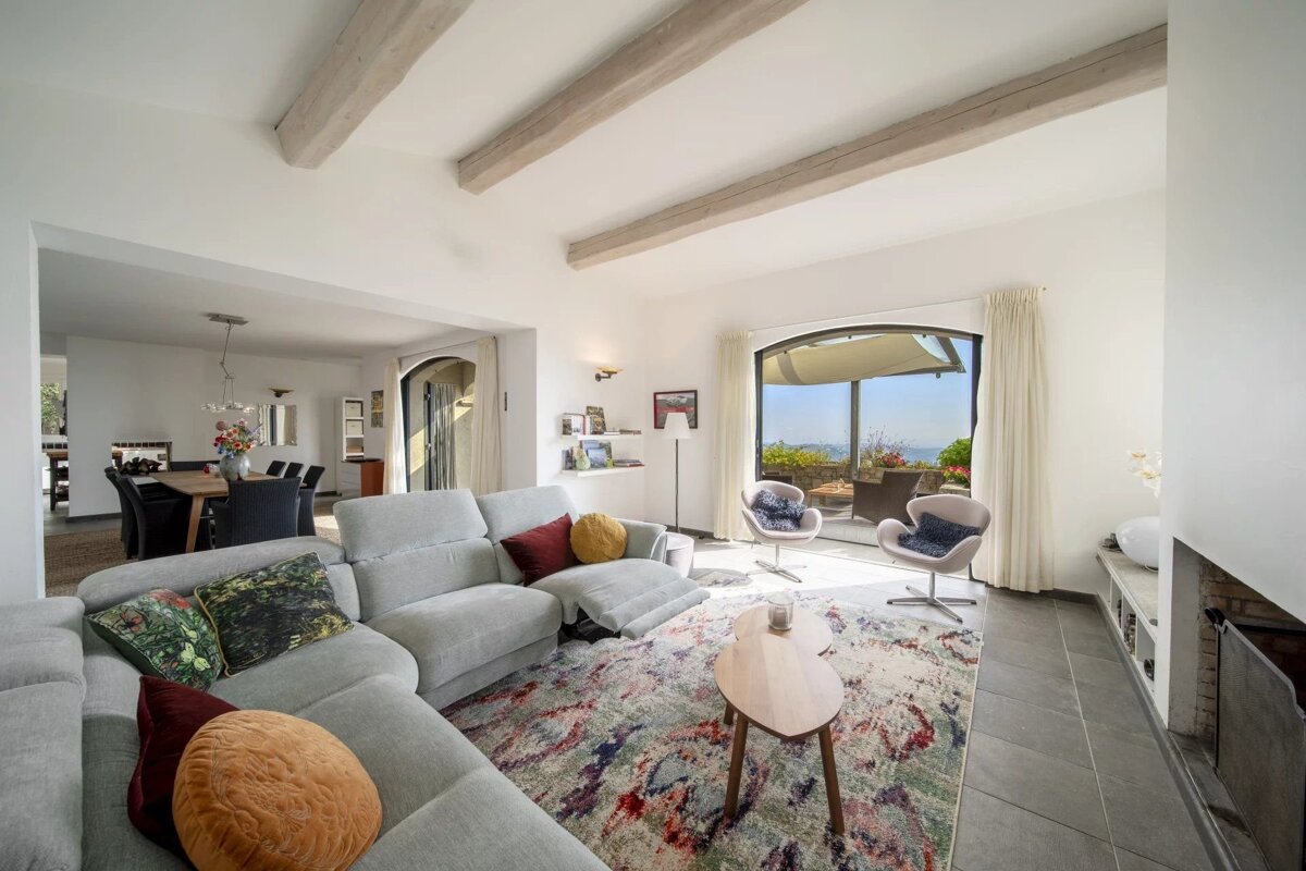 A bright, open-plan living room with a large grey sectional sofa, colorful rug, exposed beams, and a dining area, leading to a terrace with scenic views.