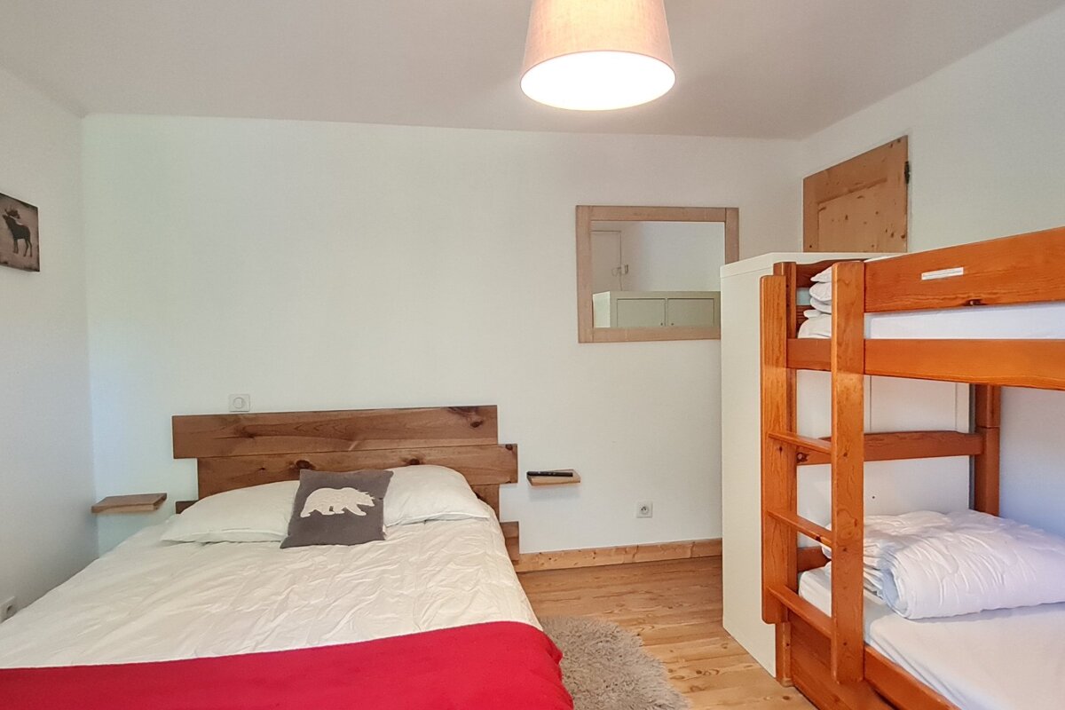 A cozy bedroom featuring a double bed with a wooden headboard and a bear pillow, plus a wooden bunk bed. White walls, wooden floors, and a mirror complete the space.