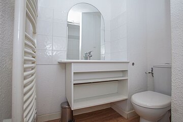 A minimalist white bathroom with an arched mirror above the sink, a toilet, and a towel warmer, set on a light wooden floor.