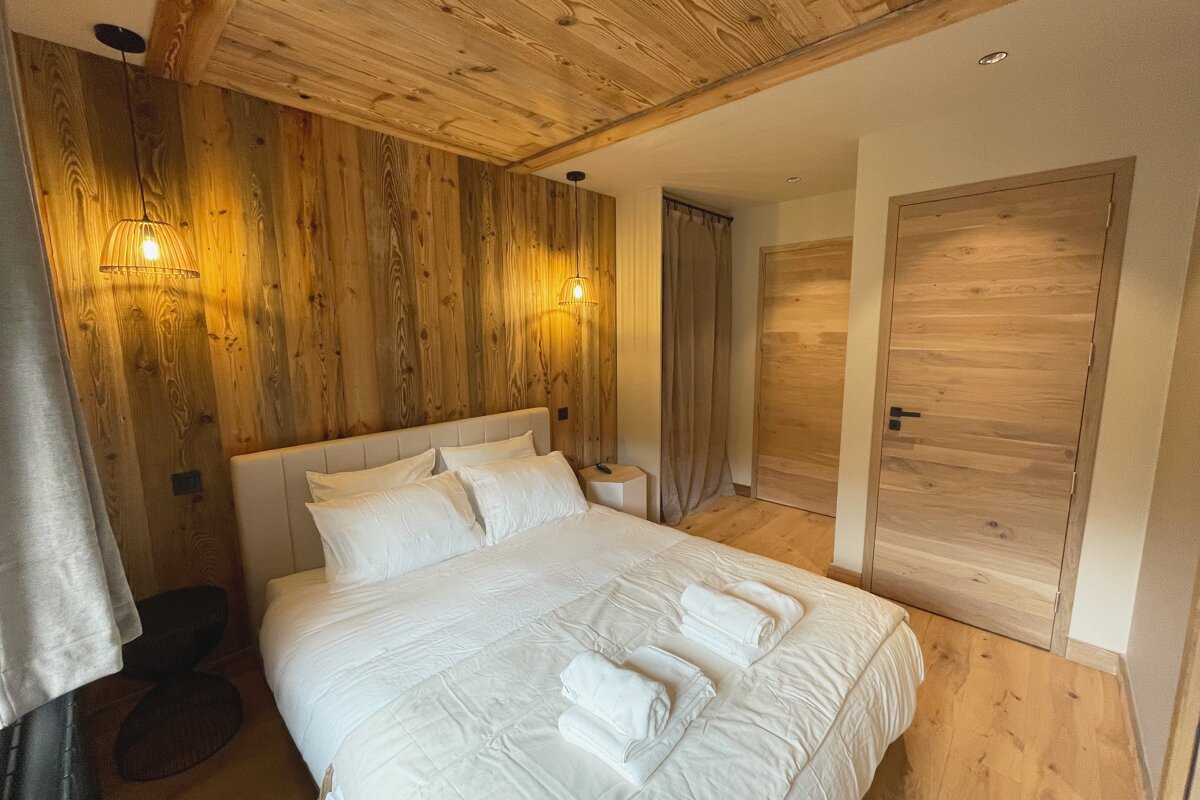 A cozy, rustic-chic bedroom features prominent wood-paneled walls and ceiling, a large bed with crisp white linens, and modern pendant lights.