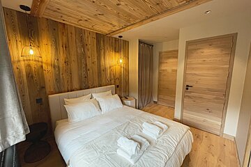 A cozy, rustic-chic bedroom features prominent wood-paneled walls and ceiling, a large bed with crisp white linens, and modern pendant lights.