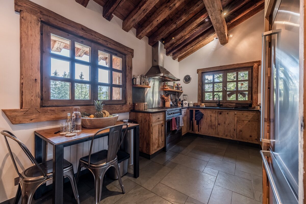 A cozy, rustic kitchen featuring exposed wood beams, windows with forest views, and a blend of natural wood cabinets with modern stainless steel appliances.