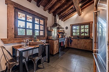 A cozy, rustic kitchen featuring exposed wood beams, windows with forest views, and a blend of natural wood cabinets with modern stainless steel appliances.