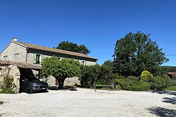 A stone house with green shutters, a car parked under an archway, and a gravel driveway surrounded by lush trees under a clear blue sky.