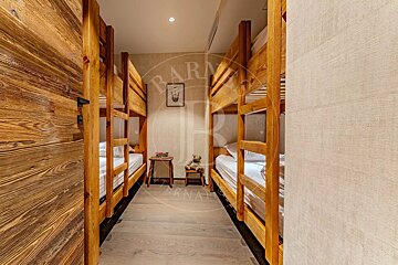 A cozy room with rustic wooden bunk beds on both sides, light wood floors, and a small teddy bear on a stool at the far end.