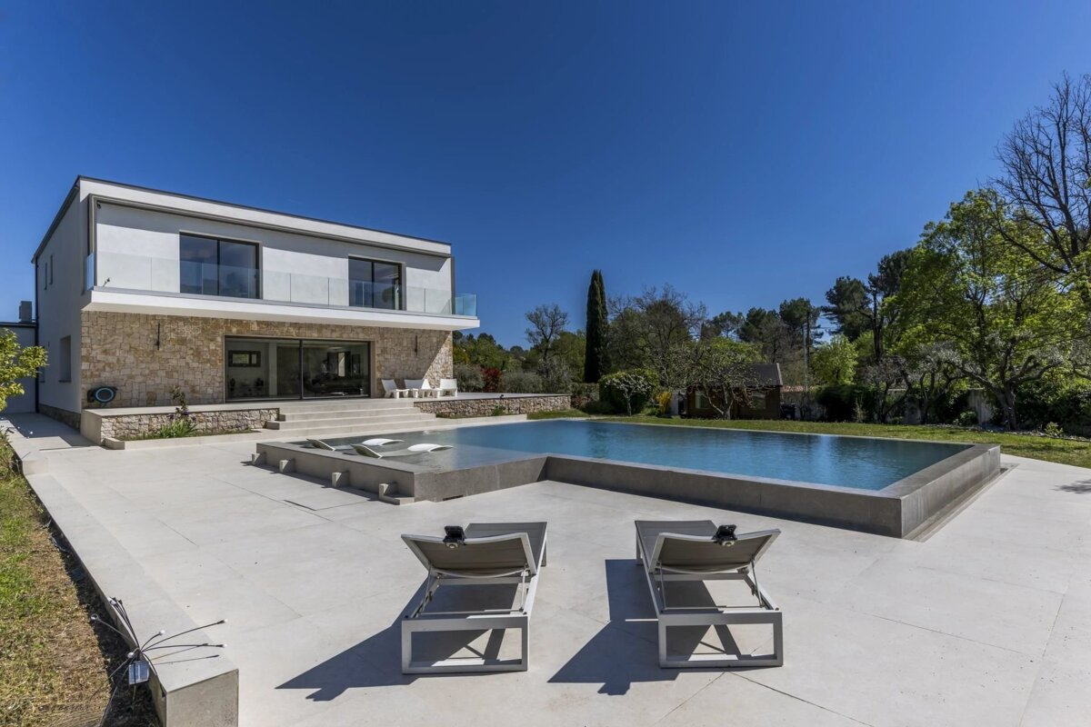 Sleek modern house with stone facade, large pool, two sun loungers on a patio, and lush greenery under a clear blue sky.