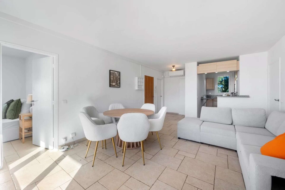 Bright, open-plan living/dining room with a grey sectional, round table & white chairs. A small kitchen is visible in the background.