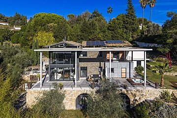 A modern, glass and stone house on a green hillside under a blue sky, featuring solar panels and a hot tub, nestled among lush trees.