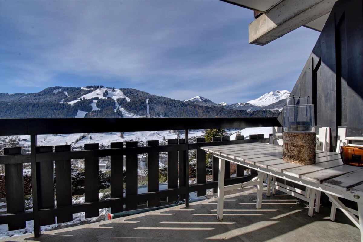 View from a balcony with a wooden table, overlooking snow-covered mountains and ski slopes under a blue sky.