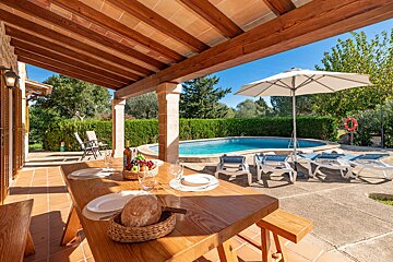 Sunny covered patio with a wooden dining table, set with bread and fruit, overlooking a clear blue swimming pool surrounded by loungers and green foliage.