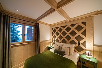 Cozy, rustic bedroom with wooden walls & ceiling beams. Features a green bed, diamond headboard, and a window overlooking a snowy landscape.