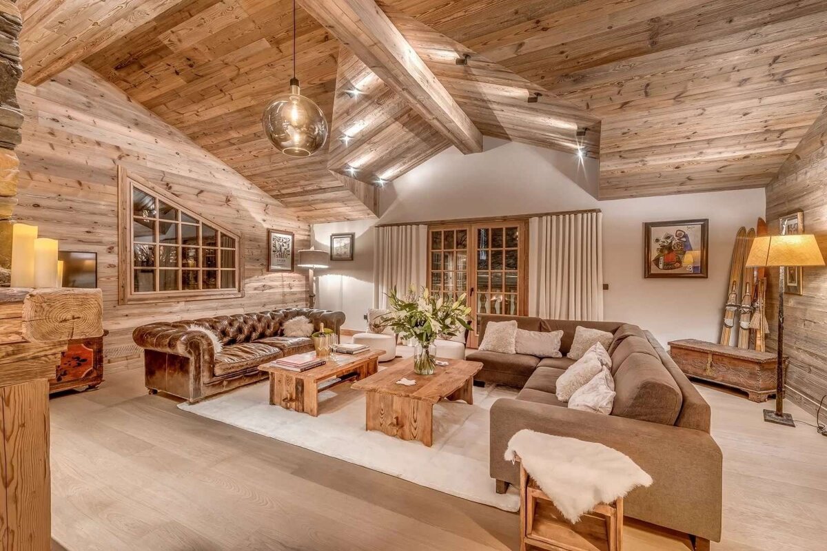 A cozy chalet living room with warm wood paneling, exposed beams, comfortable sofas, rustic tables, and inviting ambient lighting.