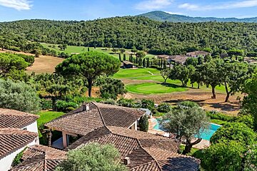 Aerial view of terracotta-roofed houses with a pool, overlooking a lush green golf course and forested hills under a blue sky.